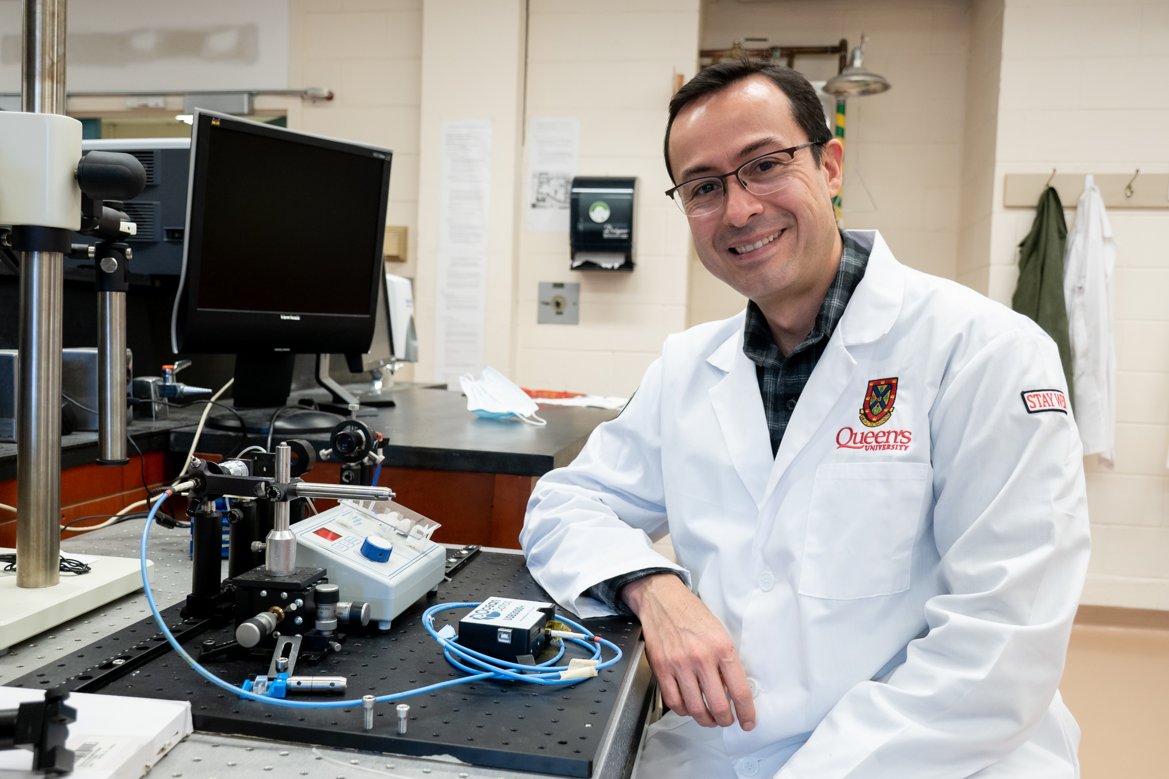 Man with glasses and lab coat sitting in a lab, he is smiling at camera