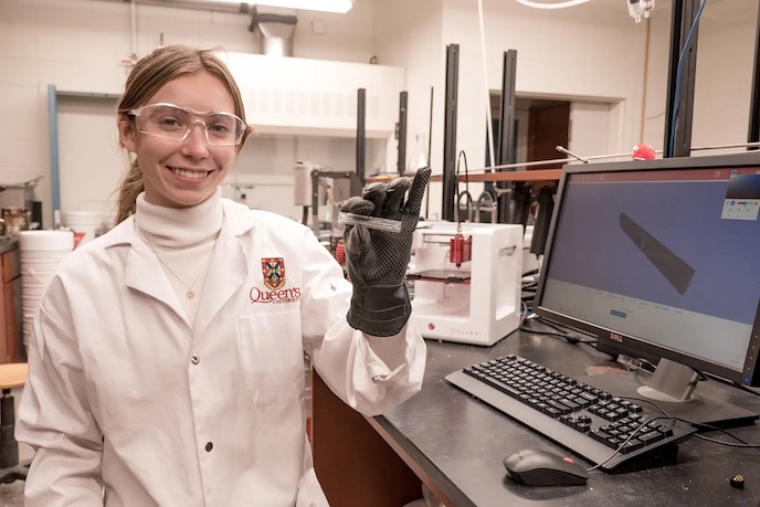 Smiling student in a lab coat and safety goggles holding a small component in a laboratory.