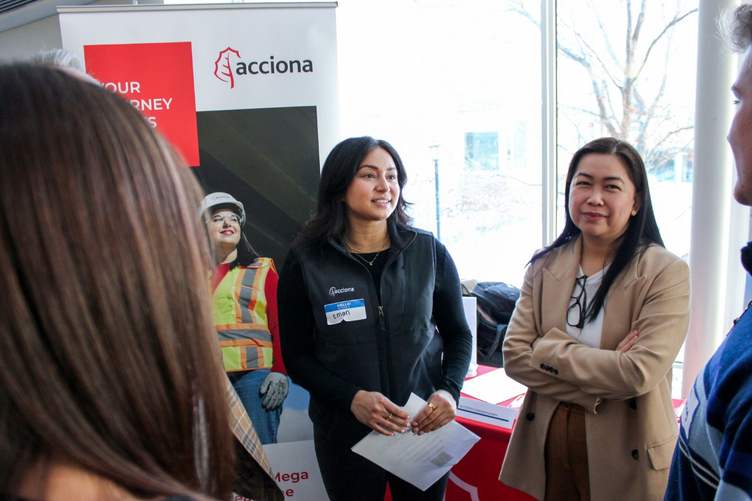Two women stand in front of a trade show booth speaking with people