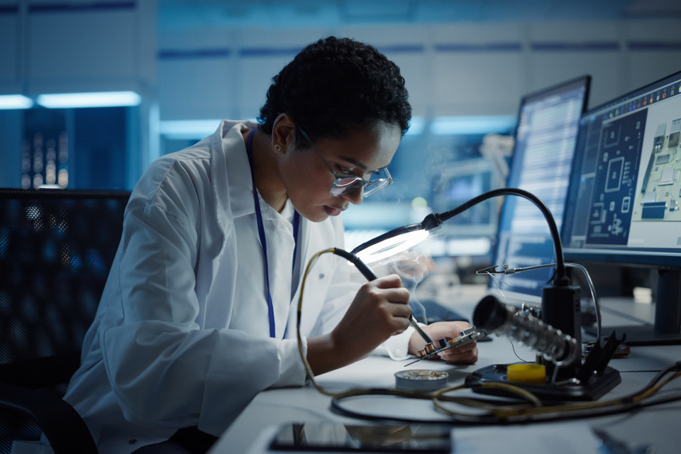 Woman engineer soldering a circuit board under a lamp at a computer-equipped lab workstation
