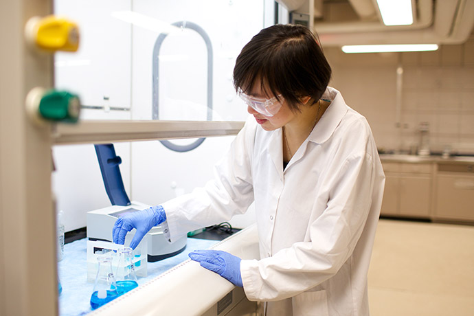 Researcher wearing safety goggles and gloves working with beakers and lab equipment at a laboratory bench.