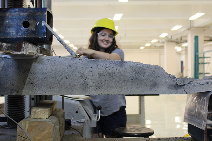 Student wearing a hard hat and safety glasses operating a concrete testing machine in a lab.