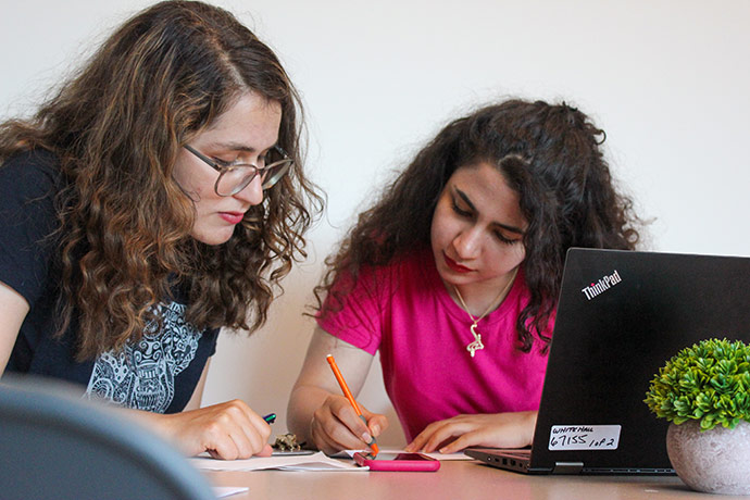 Two women studying together at a table, writing notes beside a laptop.