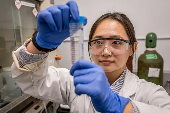 Researcher wearing safety goggles and blue gloves examining a test tube in a laboratory.