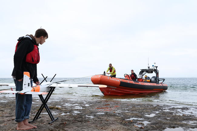 Young white man standing on a beach looking at a small model plane. There is a dingy nearby with people. 