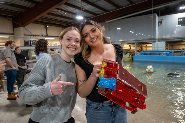 Two young women are showing off their model house. They are smiling at the camera proudly.