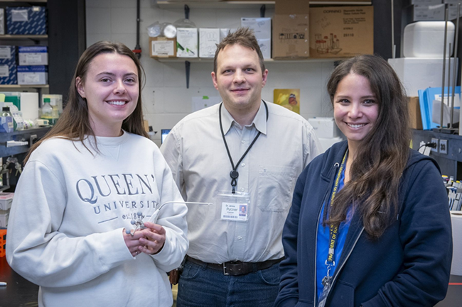 Three people are standing in a lab, looking at the camera and smiling. 