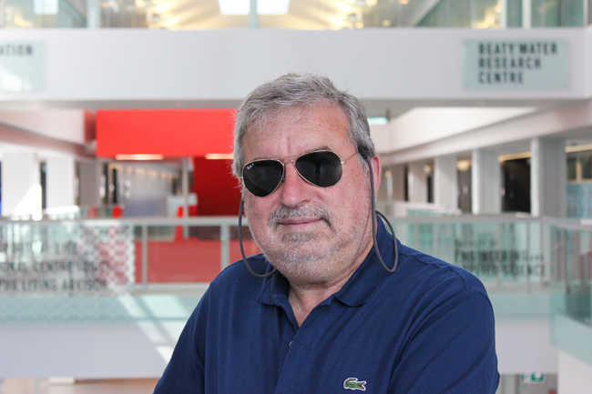 White man with grey hair standing in a modern building. He is smiling at camera and wearing aviator glasses