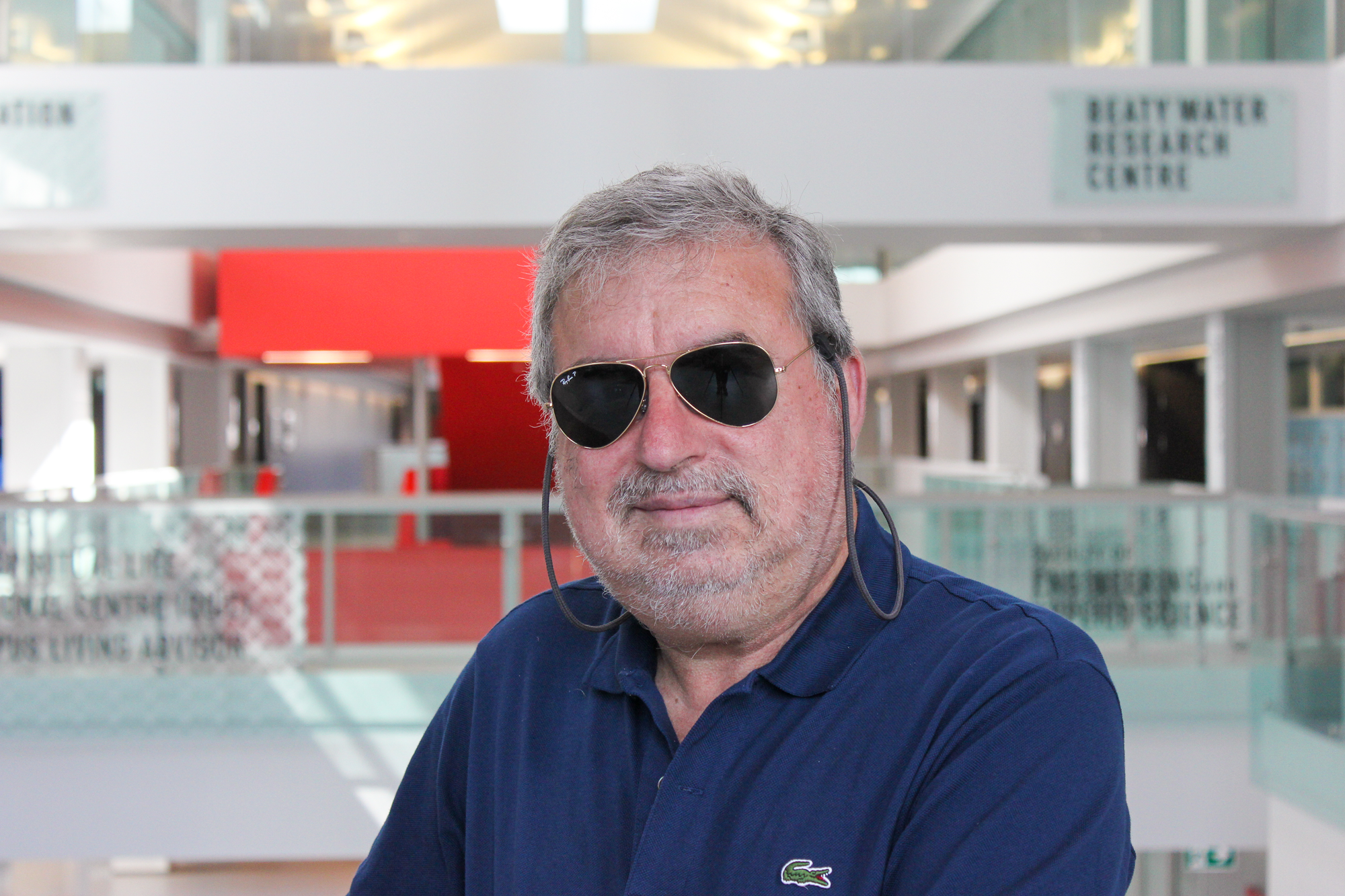 White man with grey hair in a modern building. He is smiling at the camera. He is wearing aviator glasses.